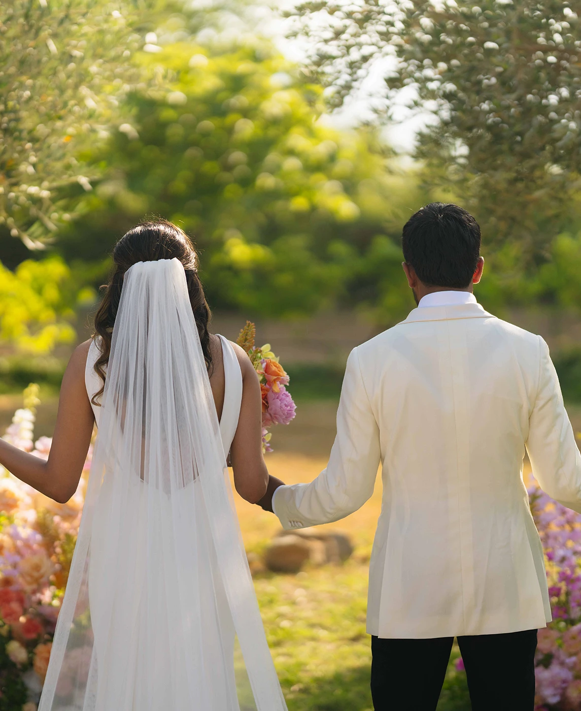 Flottie wedding decor and organisation: bride and groom seen from behind walking outdoors through a sunlit garden surrounded by abundant floral styling for the Gathered in Joy project