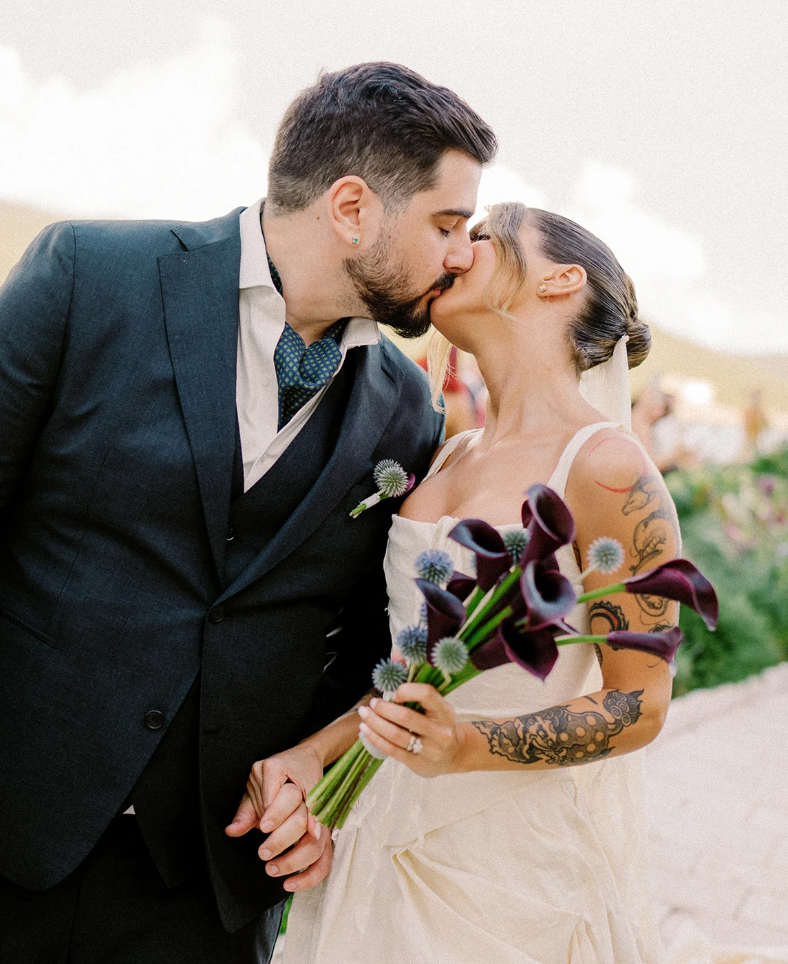 Flottie wedding decor and organisation: romantic couple kissing outdoors with the groom in a dark suit and the bride holding a custom bouquet of dark calla lilies for the Mulberry Tide wedding project