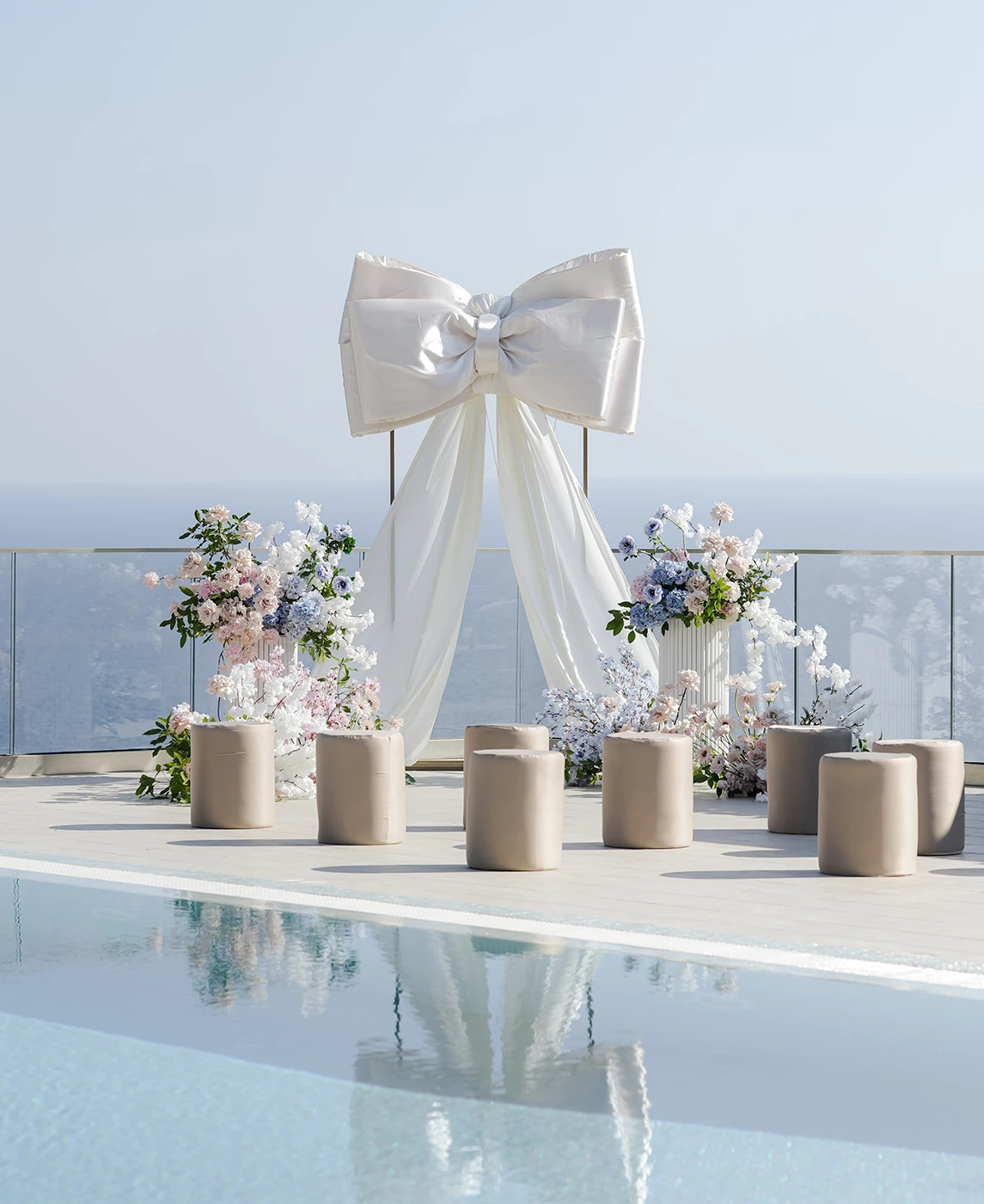 Flottie wedding decor and organisation: modern poolside wedding ceremony setup featuring a large white decorative bow altar, cylindrical seating, and a champagne tower for the A Toast To Me project
