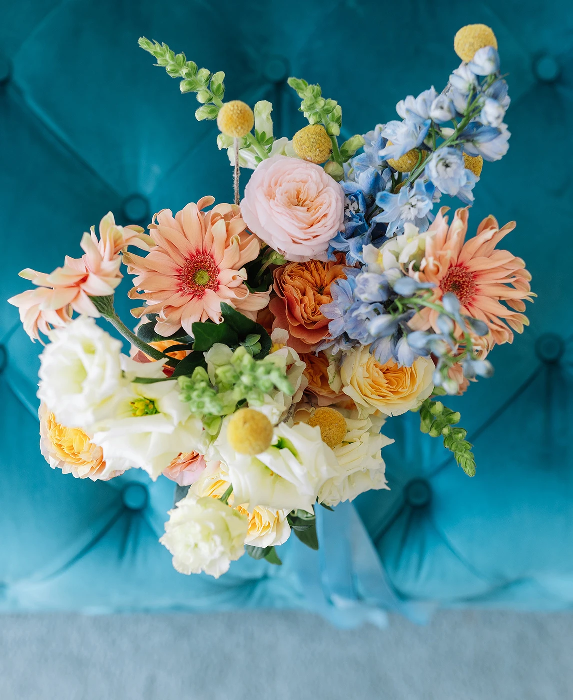 Flottie wedding decor and organisation: vibrant floral bouquet featuring pastel roses, gerberas, and blue accents resting against a tufted turquoise velvet background for the Heritage of Love project