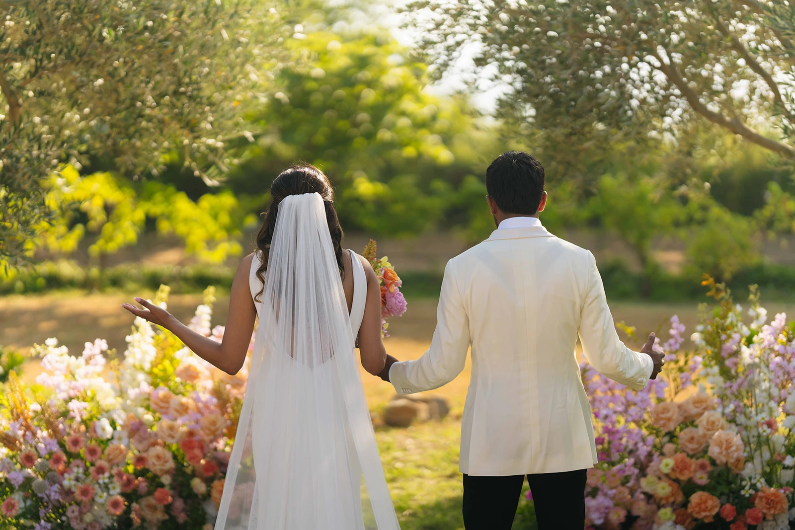 Flottie wedding decor and organisation: bride and groom seen from behind walking outdoors through a sunlit garden surrounded by abundant floral styling for the Gathered in Joy project