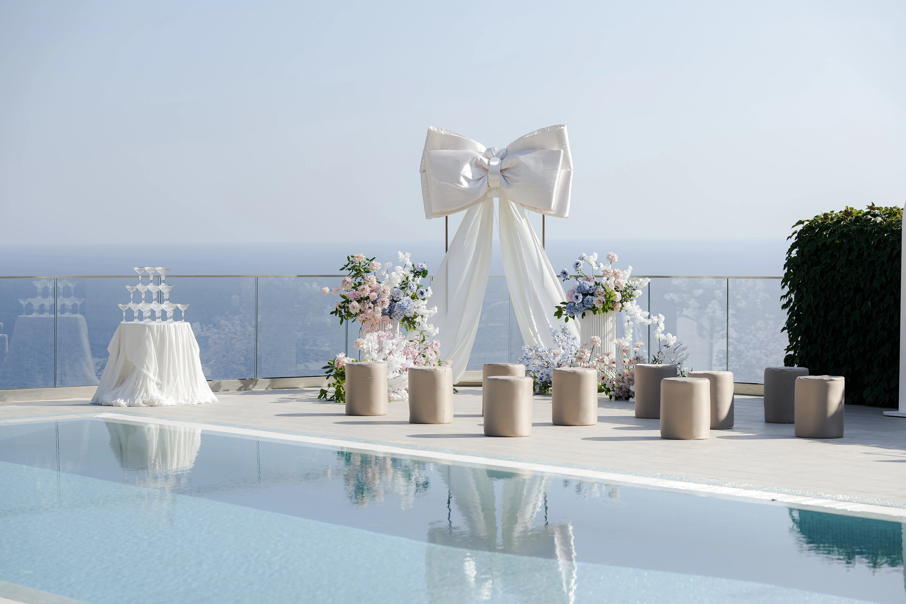Flottie wedding decor and organisation: modern poolside wedding ceremony setup featuring a large white decorative bow altar, cylindrical seating, and a champagne tower for the A Toast To Me project
