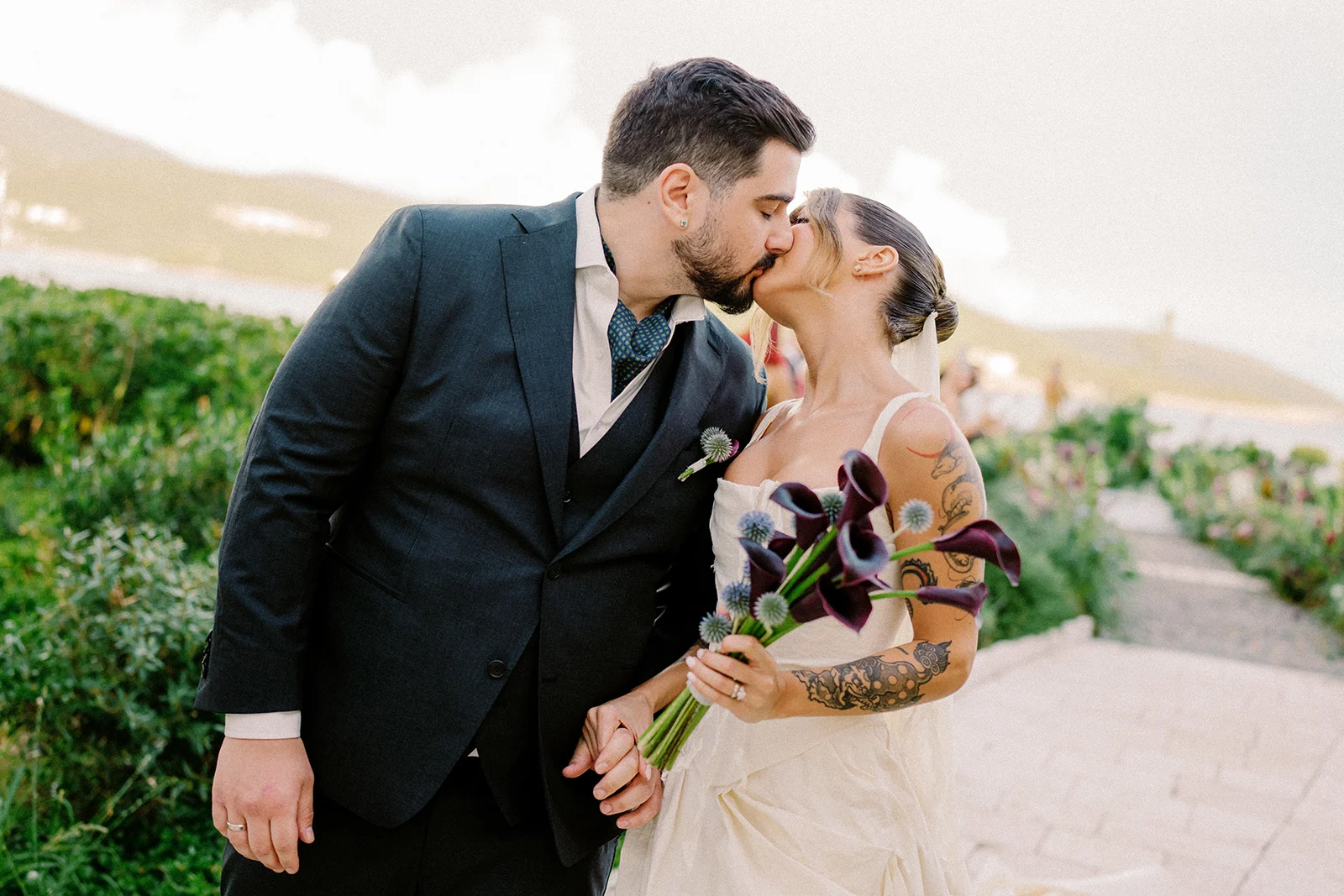 Flottie wedding decor and organisation: romantic couple kissing outdoors with the groom in a dark suit and the bride holding a custom bouquet of dark calla lilies for the Mulberry Tide wedding project