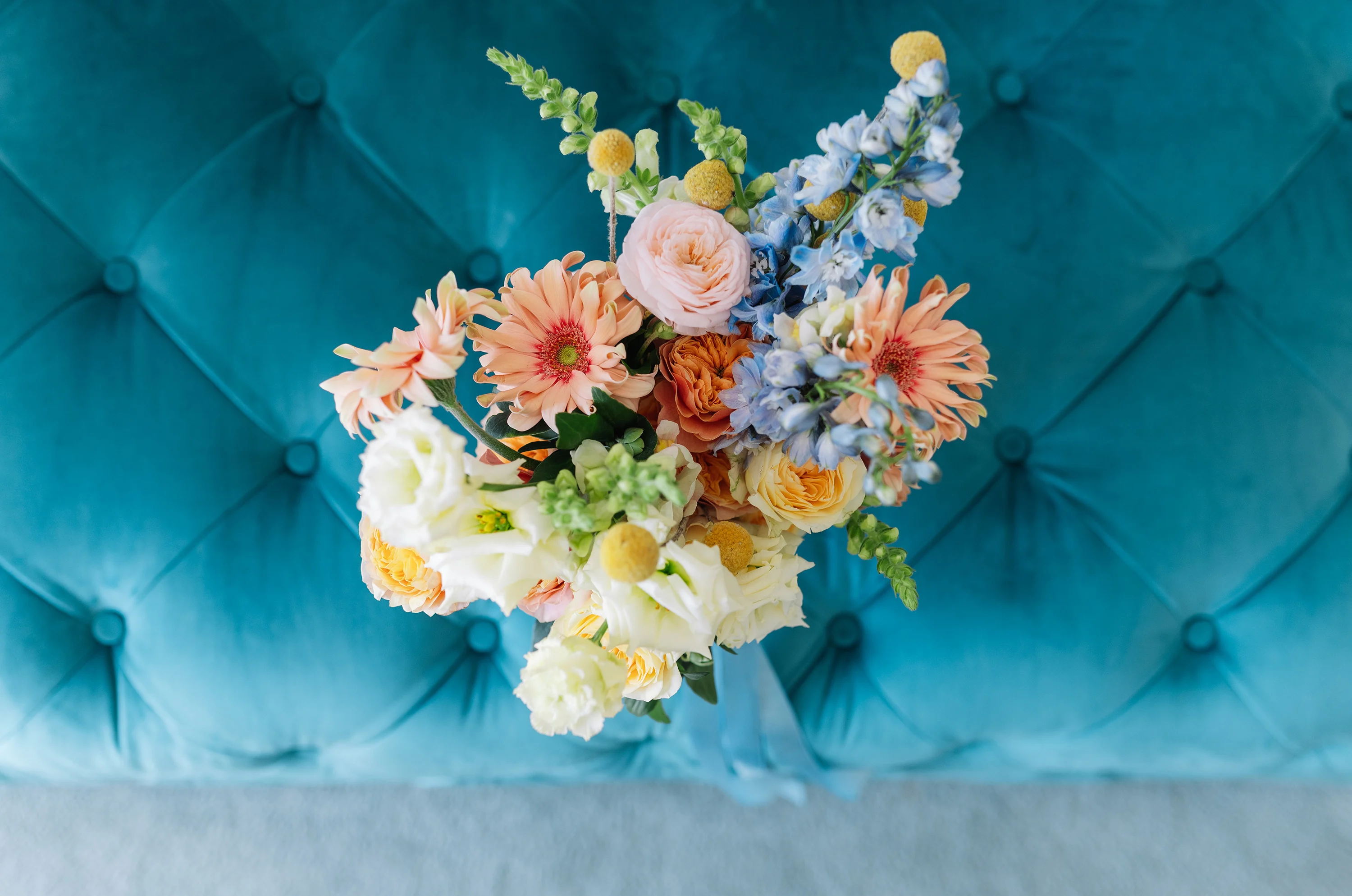 Flottie wedding decor and organisation: vibrant floral bouquet featuring pastel roses, gerberas, and blue accents resting against a tufted turquoise velvet background for the Heritage of Love project